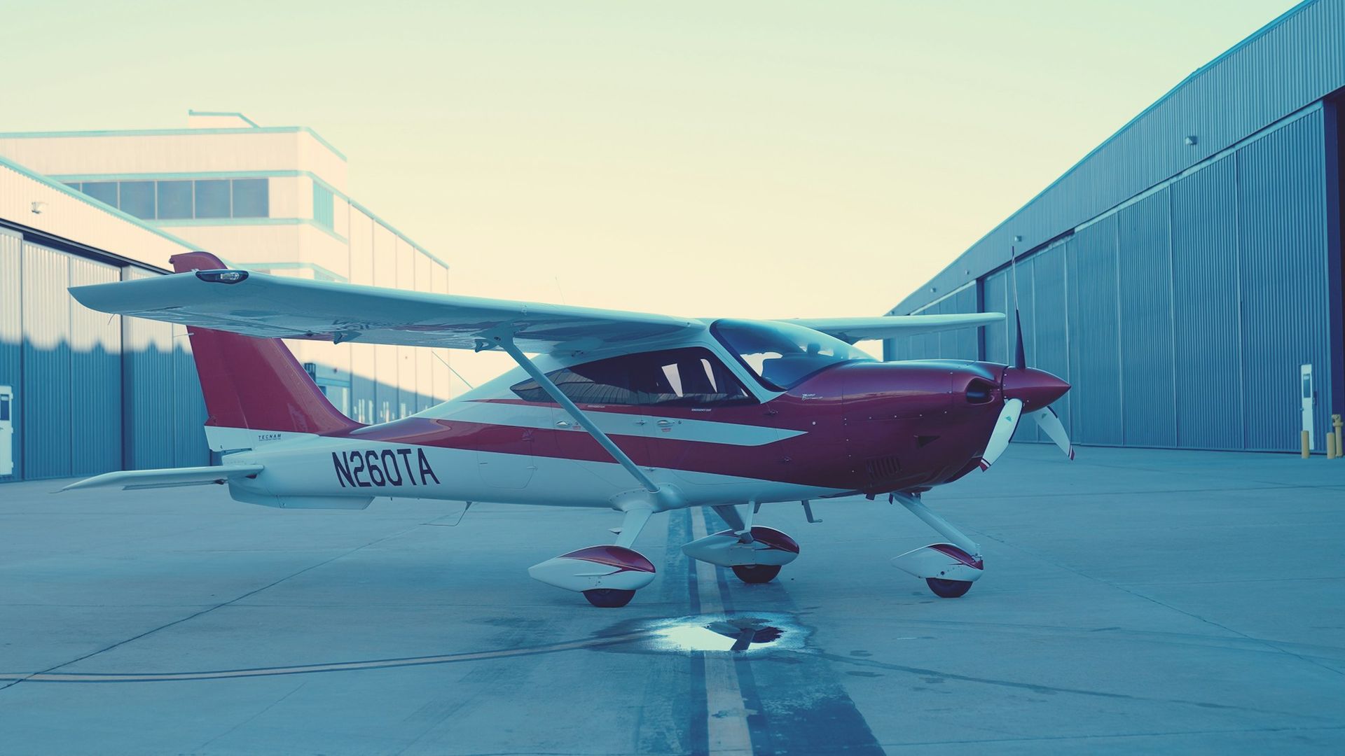 Un avion léger rouge et blanc stationné sur le tarmac, devant ses hangars